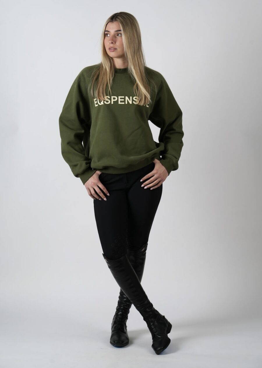 Woman in green sweatshirt and black pants posing against a plain background.