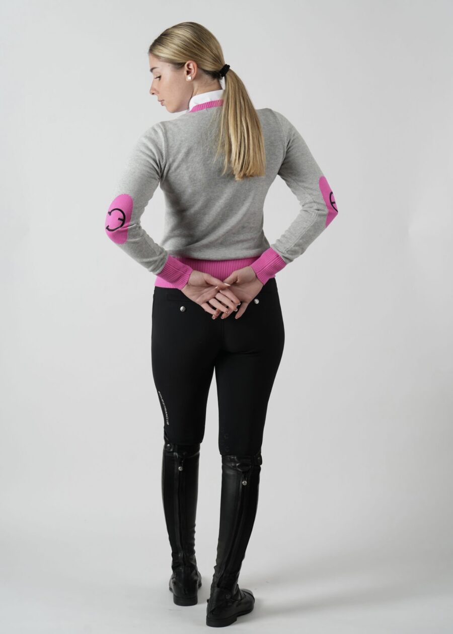 Woman in stylish equestrian outfit with pink elbow patches stands with hands behind back.