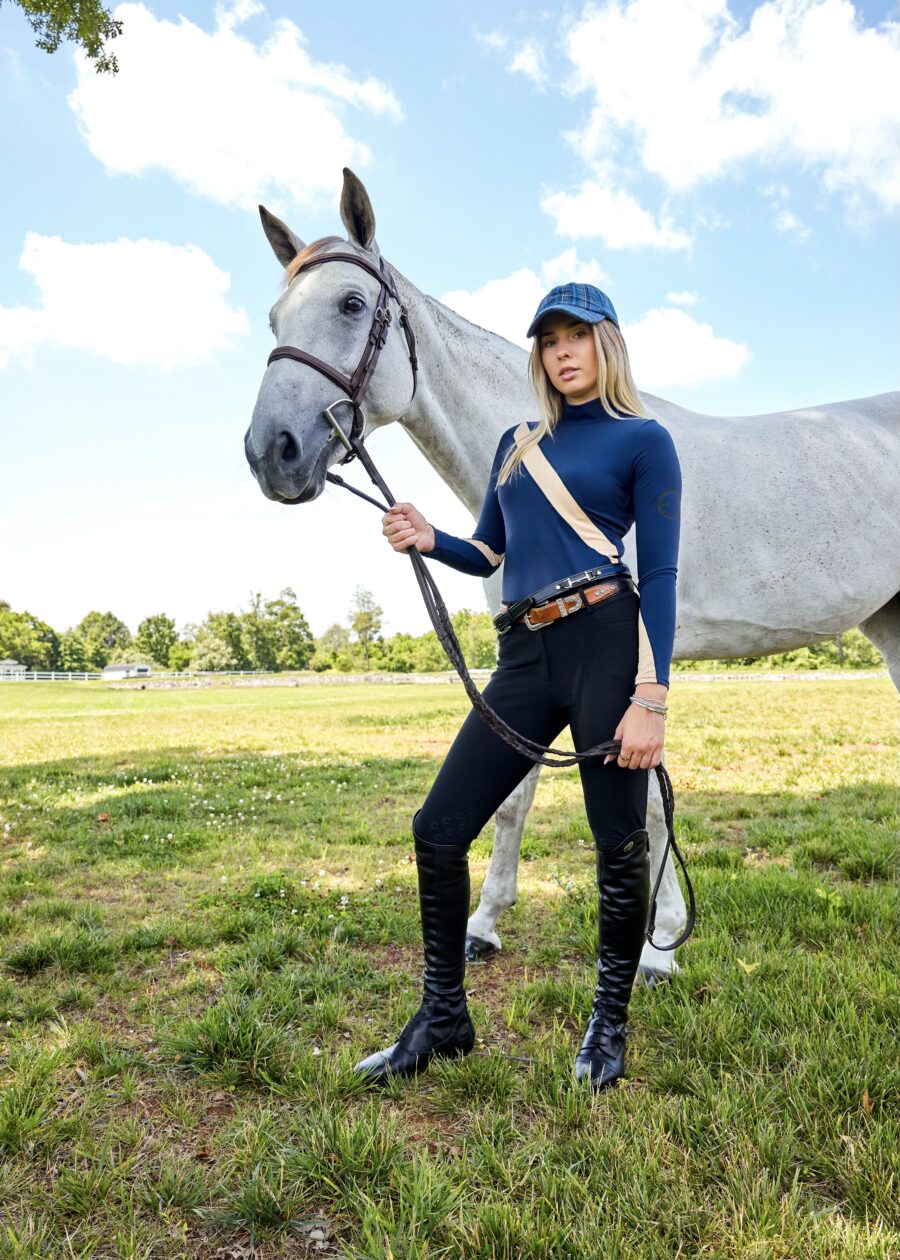 A woman in equestrian attire holding a white horse in a grassy field.
