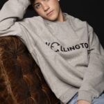 Young man wearing a gray Wellington sweatshirt sitting on a brown leather couch.