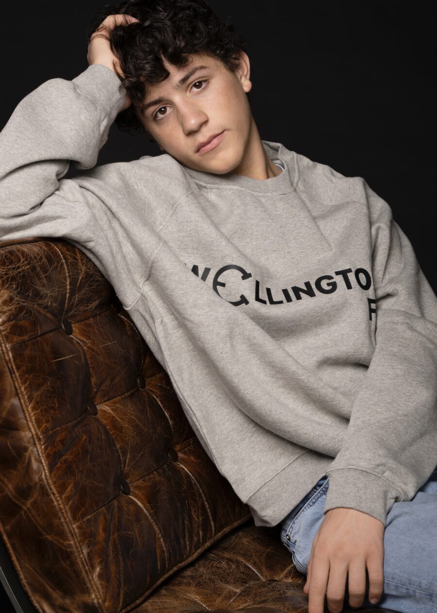 Young man wearing a gray Wellington sweatshirt sitting on a brown leather couch.