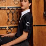 Young man sitting in a tack room with horse bridles hanging behind him.