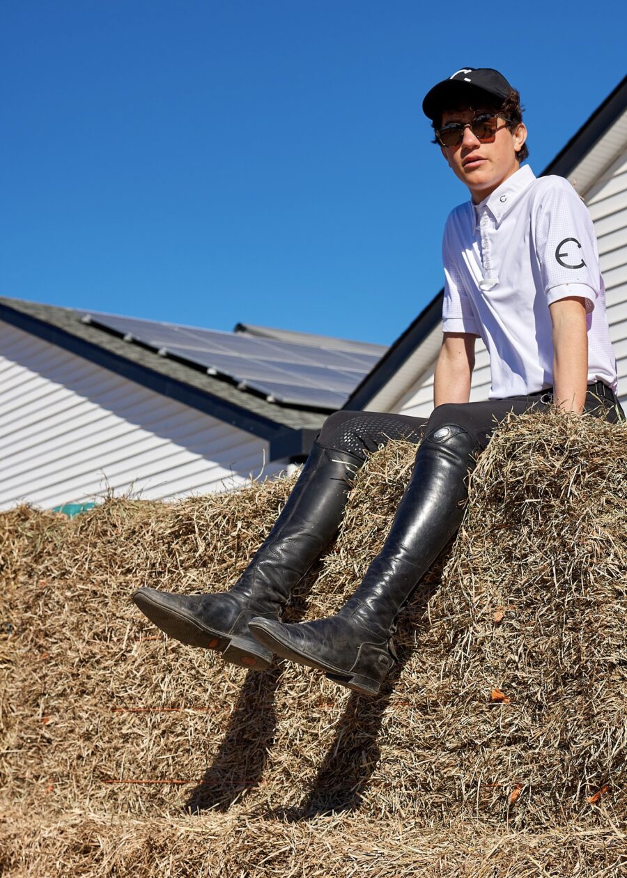 Person in white shirt and sunglasses sitting on haystack wearing shiny black boots.