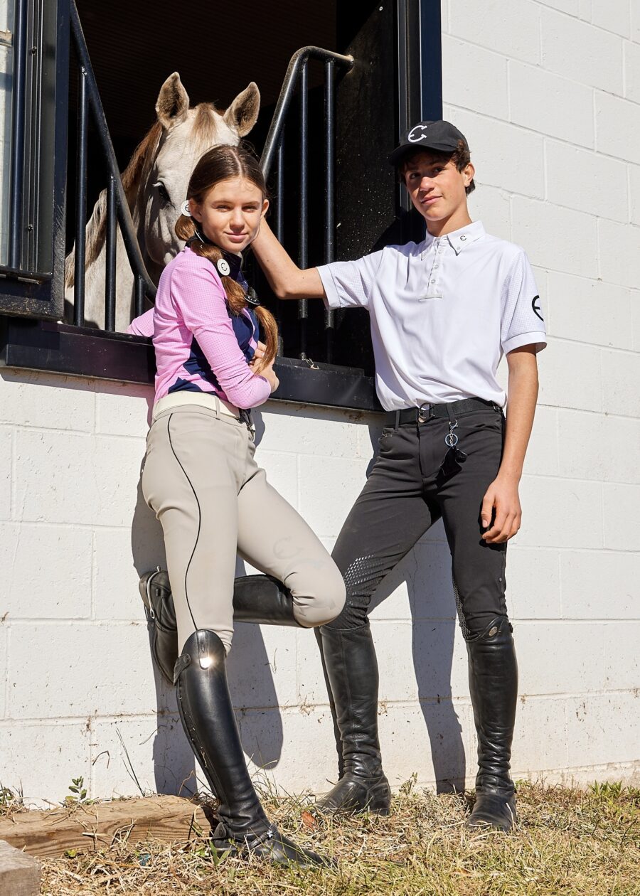 Two young equestrians posing in riding gear against a white wall.